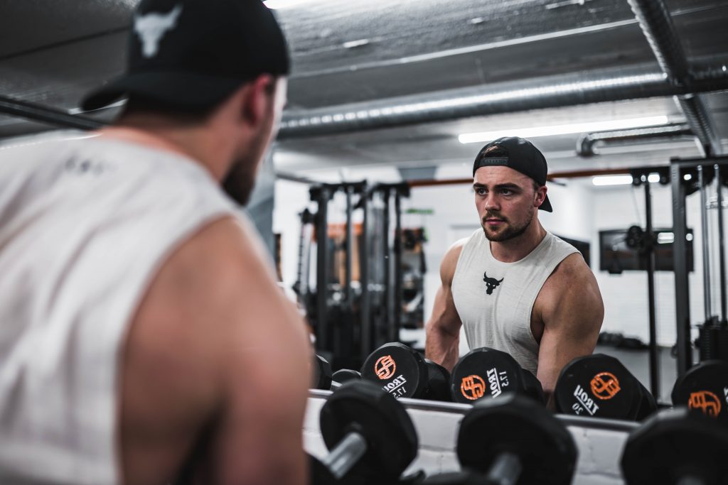 Man in baseball cap and vest using gym mirror for lifting free weights.