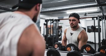 Man in baseball cap and vest using gym mirror for lifting free weights.